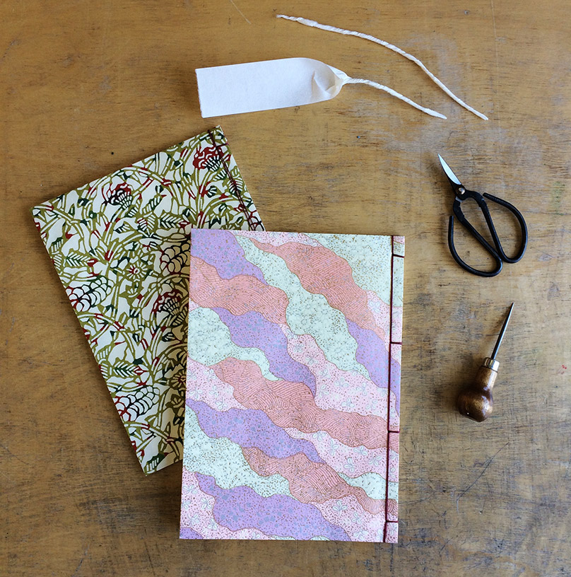 Two Japanese 4-Hole bindings laying on a wooden table with some bookbinding tools.