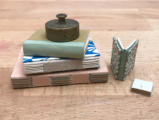 Stack of miniature bindings sitting on wooden table with weight on top.