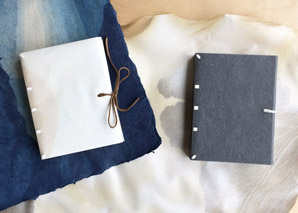 Two examples of the Limp Vellum Binding, one made with vellum and one made with handmade paper. Sitting ontop of a piece of handmade paper and vellum respectively.